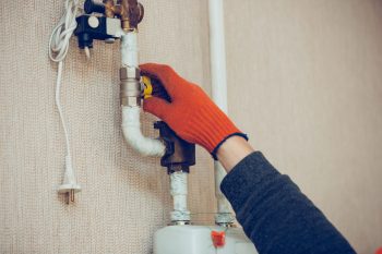 Person in orange gloves adjusting a gas valve on plumbing pipes, illustrating home maintenance for plumbing and gas fitting services.